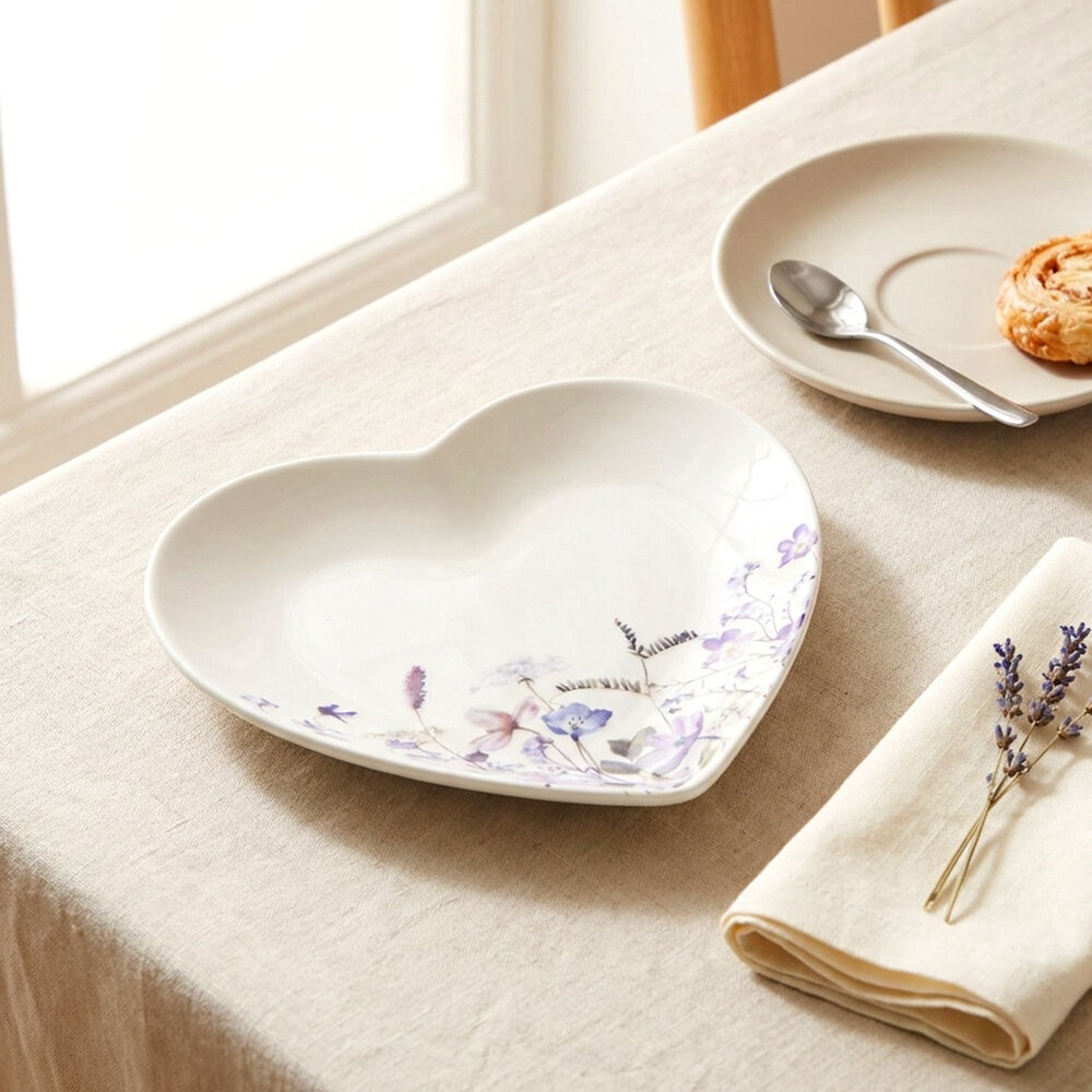 Na stole znajduje się porcelanowy talerz w kształcie serca z delikatnym kwiatowym wzorem. Obok leży beżowy talerz z łyżeczką i ciastkiem oraz złożona serwetka z gałązką lawendy.