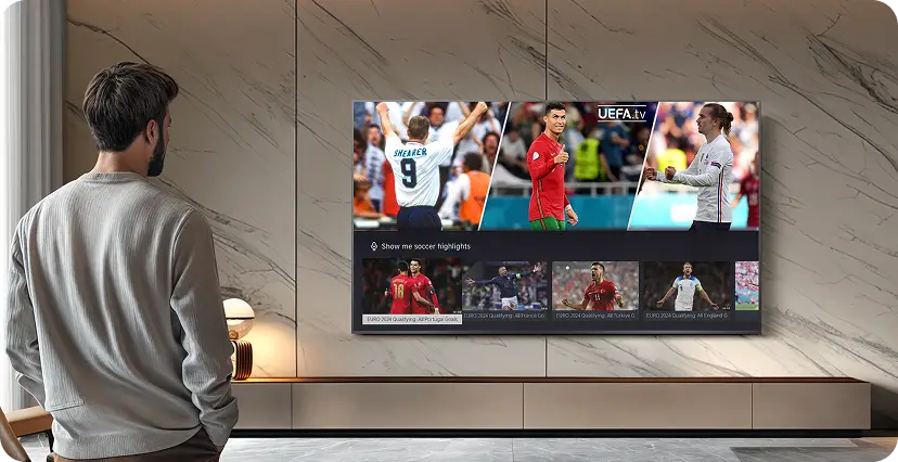 Mężczyzna stoi przed dużym telewizorem zamontowanym na ścianie, na którym wyświetlane są obrazy z meczu piłki nożnej oraz logo UEFA.tv. W tle widać jasną, marmurową ścianę i minimalistyczne meble.