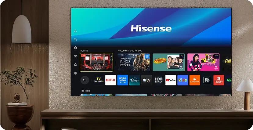 Telewizor wyświetla ekran główny z logo Hisense na górze i aplikacjami takimi jak Netflix, Prime Video, Disney+, HBO Max, i Apple TV. W tle widoczna jest część pomieszczenia z lampą i rośliną na półce.
