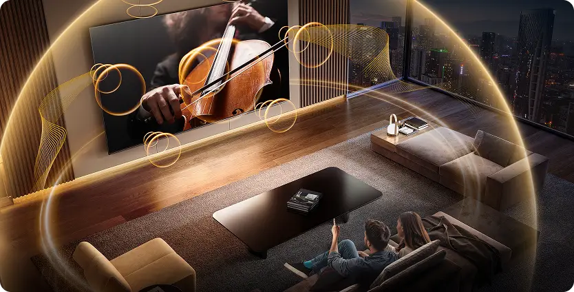 W przestronnym salonie znajduje się duży telewizor wyświetlający obraz osoby grającej na instrumencie smyczkowym. Wokół telewizora widoczne są efekty świetlne, a na kanapie siedzą dwie osoby.