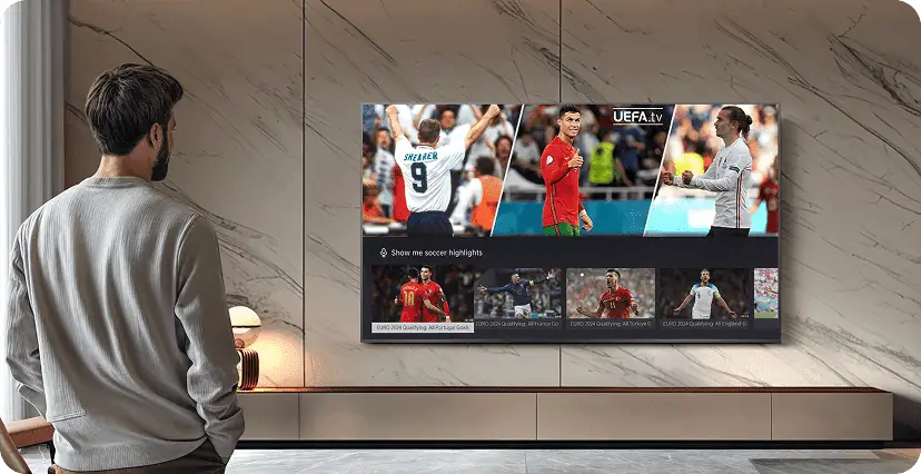 Na ścianie wisi duży telewizor, na którym wyświetlane są obrazy z meczu piłki nożnej oraz napis \'UEFA.tv\'. Przed telewizorem stoi osoba, patrząc na ekran.