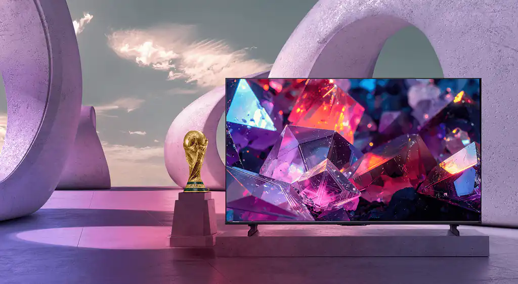 Telewizor Hisense E7 Pro z kolorową grafiką kryształów obok statuetki FIFA World Cup na tle betonowych, owalnych łuków.