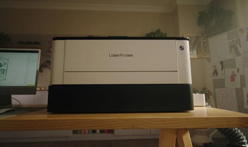 Na drewnianym stole stoi urządzenie z napisem \'LaserPecker\'. W tle widoczny jest monitor oraz ściana z rysunkami i roślinami.