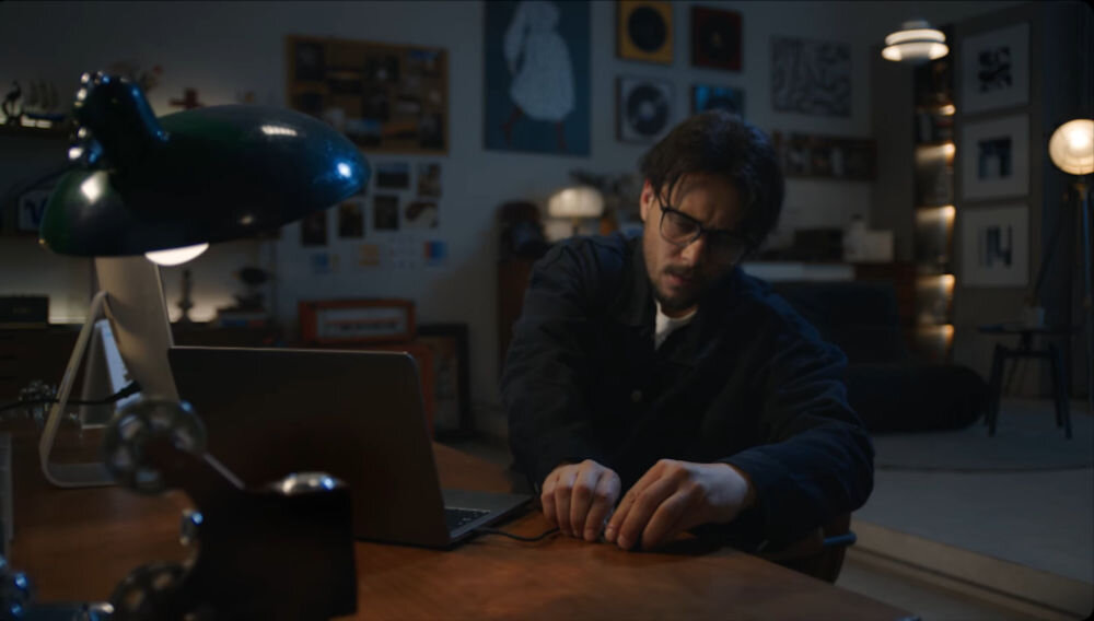 System bezprzewodowy HOLLYLAND Lark Max 2 Ultimate Combo Mężczyzna w okularach pracuje przy komputerze w słabo oświetlonym pokoju system timecode synchronizacja materiału wideo z kamery z nagraniem audio wyjścia 3.5 mm i UAC wyrównanie audio-wizualne