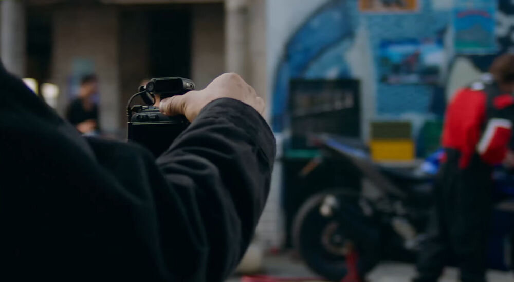 System bezprzewodowy HOLLYLAND Lark Max 2 Ultimate Combo Mężczyzna trzyma kamerę i nagrywa mężczyznę ubranego w czerwono-białe ubrania stojący przy motocyklu transmisja dźwięku wysokiej jakości do 340 m swobodne poruszanie się