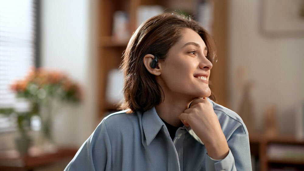 System bezprzewodowy HOLLYLAND Lark Max 2 Ultimate Combo Kobieta ze słuchawką OWS i mikrofonem MAX 2, opierająca brodę na dłoni, ubrana w niebieską koszulę słuchawki OWS otwarta konstrukcja regulowane zaczepy dopasowanie