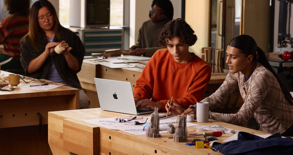 Laptop APPLE MacBook Pro 2026 Dwoje młodych projektantów pracujących przy drewnianym stole warsztatowym nad planami architektonicznymi i modelami 3D z wykorzystaniem laptopa MacBook.
