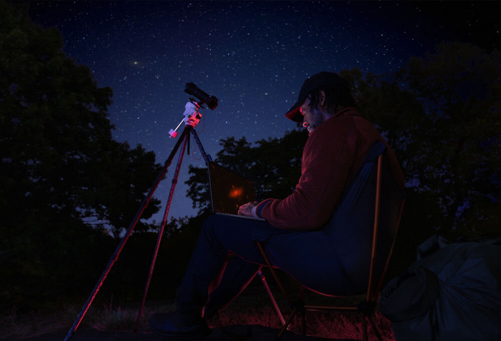 Laptop APPLE MacBook Pro 2026 Mężczyzna siedzący na kempingu w nocy, używający laptopa MacBook obok teleskopu na statywie pod rozgwieżdżonym niebem.
