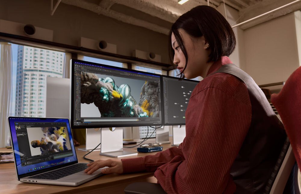 Laptop APPLE MacBook Pro 2026 Kobieta pracująca w profesjonalnym studio przy biurku z laptopem i dwoma dużymi monitorami zewnętrznymi, na których widoczne są złożone modele 3D i struktury węzłowe.