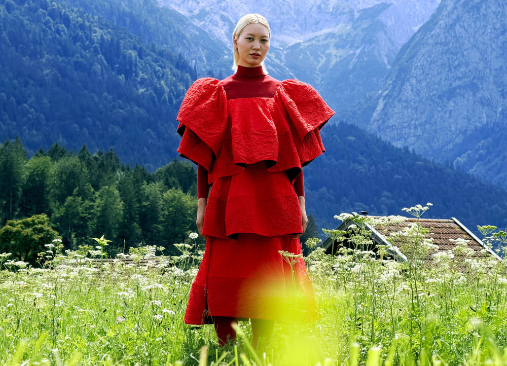 Monitor APPLE Studio Display XDR Portret kobiety w awangardowej czerwonej sukni z falbanami, stojącej na łące na tle wysokich, zamglonych gór.