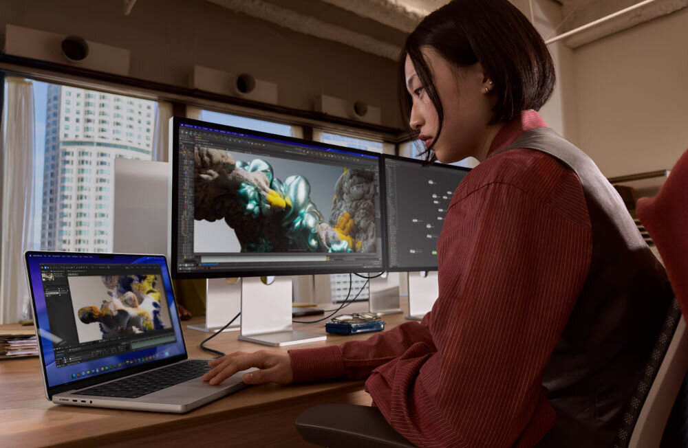 Monitor APPLE Studio Display XDR Kobieta pracująca przy biurku z laptopem podłączonym do dwóch zewnętrznych monitorów wyświetlających zaawansowane projekty graficzne 3D w profesjonalnym studiu.