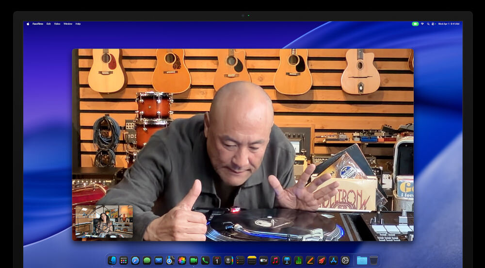Monitor APPLE Studio Display XDR Okno aplikacji FaceTime na pulpicie macOS, przedstawiające mężczyznę w studiu muzycznym z kolekcją gitar na ścianie, wykonującego gest kciuka w górę nad gramofonem.