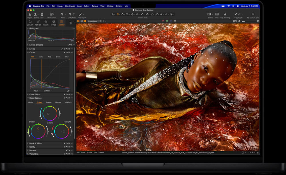 Laptop APPLE MacBook Pro 2026 Kobieta w kolorowym swetrze pracująca przy biurku nad montażem wideo Community Garden na komputerze Mac z podłączonym profesjonalnym mikrofonem i oświetleniem.