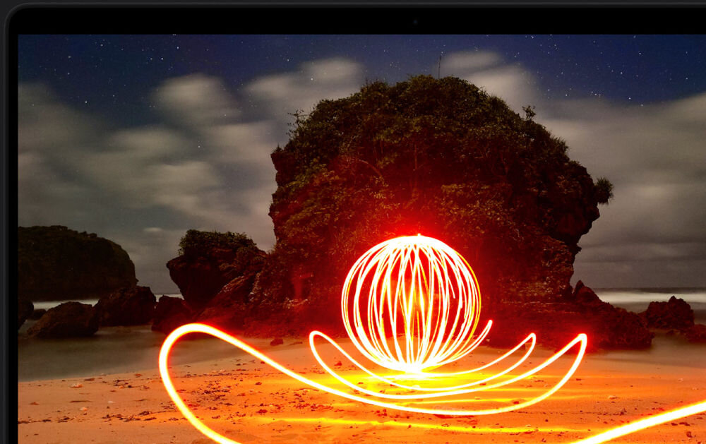 Laptop APPLE MacBook Pro 2026 Zbliżenie na ekran laptopa wyświetlający nocne zdjęcie plaży z artystycznym malowaniem światłem w kształcie świecącej kuli przed formacjami skalnymi.