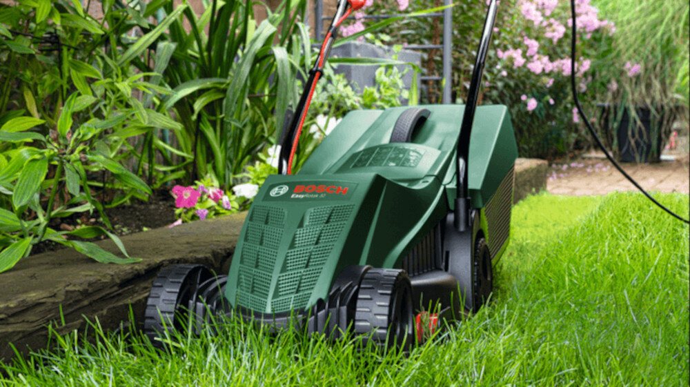 Kosiarka elektryczna BOSCH EasyRotak 32-235 1200 W 320 mm ogród zbliżenie na korpus kosiarki w tle kwiaty szczotkowy silnik mocy 1200 W z momentem obrotowym 13 Nm efektywne koszenie nawet na gęstych i wilgotnych fragmentach trawnika stała prędkość noża system ochrony przed przeciążeniem zabezpiecza urządzenie przed uszkodzeniem