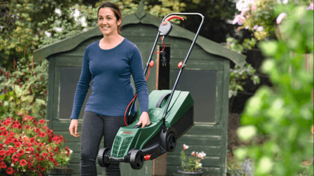 Kosiarka elektryczna BOSCH EasyRotak 32-235 1200 W 320 mm ogród kobieta idzie z kosiarka w dłoni w tle szopa kwiaty drzewa waga 10 kg zintegrowany uchwyt do przenoszenia lepsze efekty koszenia łatwe prowadzenie poziom hałasu na poziomie 92 dB intuicyjne sterowanie proste funkcje