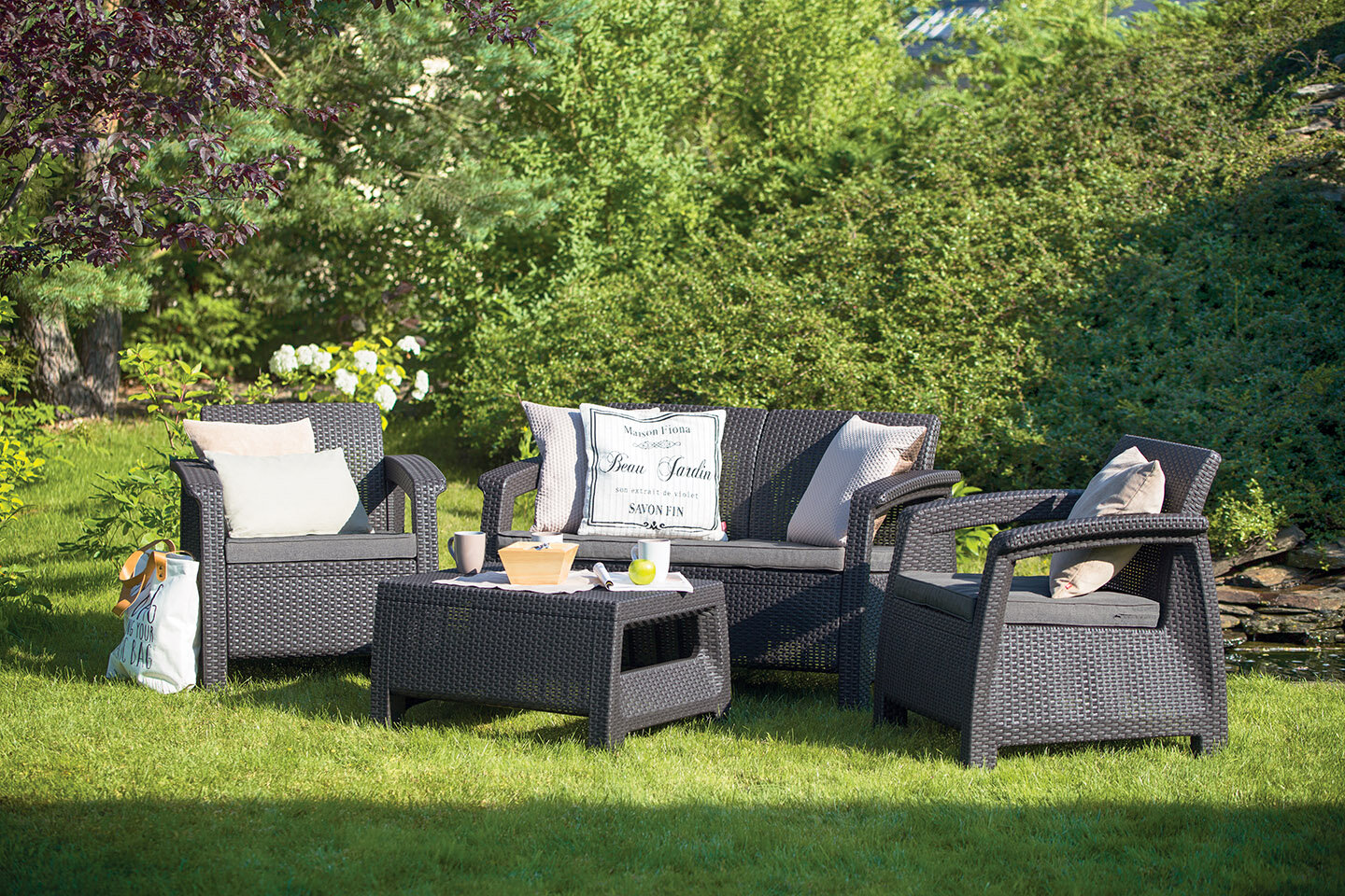 Na trawniku znajduje się zestaw mebli ogrodowych w kolorze grafitowym, składający się z dwóch foteli, sofy i stolika. Na meblach leżą poduszki, a na stoliku stoją kubki i miska.