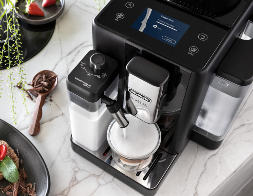 Ekspres do kawy DELONGHI Rivelia z wyświetlaczem dotykowym i dwoma wymiennymi zbiornikami na kawę, przygotowujący kawę mrożoną. Obok ekspresu znajduje się filiżanka z mleczną pianką.