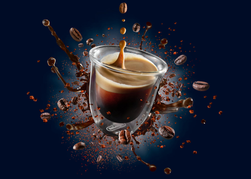 Szklanka z kawą espresso, z której rozpryskuje się kawa, otoczona unoszącymi się ziarnami kawy na ciemnym tle.