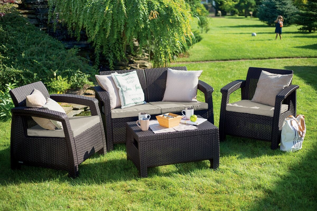 Zestaw mebli ogrodowych składający się z dwóch foteli, sofy i stolika, wykonany z brązowego materiału imitującego rattan, z beżowymi poduszkami. Na stoliku znajdują się filiżanki, miska z owocami i gazeta, a w tle widoczny jest zielony ogród.