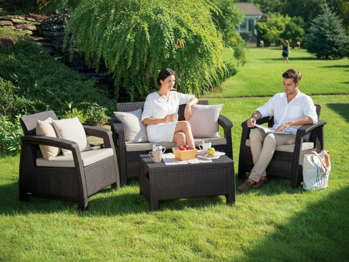 Na obrazie widoczny jest zestaw mebli ogrodowych w kolorze brązowo-beżowym, składający się z dwóch foteli, sofy i stolika, ustawionych na trawie w ogrodzie. Na stoliku znajdują się filiżanki, talerz z owocami i miska.