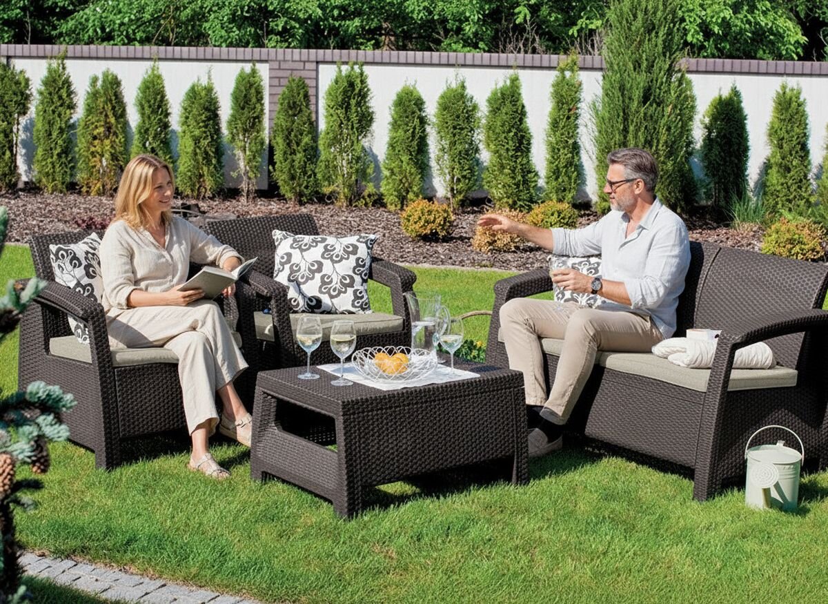 Na trawniku znajduje się zestaw mebli ogrodowych z ciemnego rattanu, składający się z dwóch foteli, sofy i stolika. Na stoliku stoją kieliszki z napojami i miska z przekąskami, a w tle widoczny jest żywopłot z iglaków.