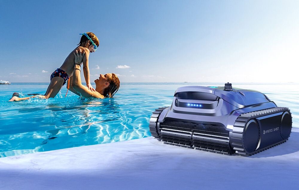 Na obrazie widać basen, w którym osoba dorosła trzyma dziecko nad wodą. Obok na brzegu znajduje się robot do czyszczenia basenów z widocznym logo \'media expert\'.