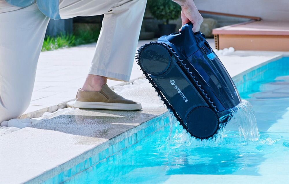 Osoba trzymająca niebieski robot do czyszczenia basenów nad krawędzią basenu, z którego wylewa się woda. Na urządzeniu widoczne jest logo \'WYBOTICS\'.
