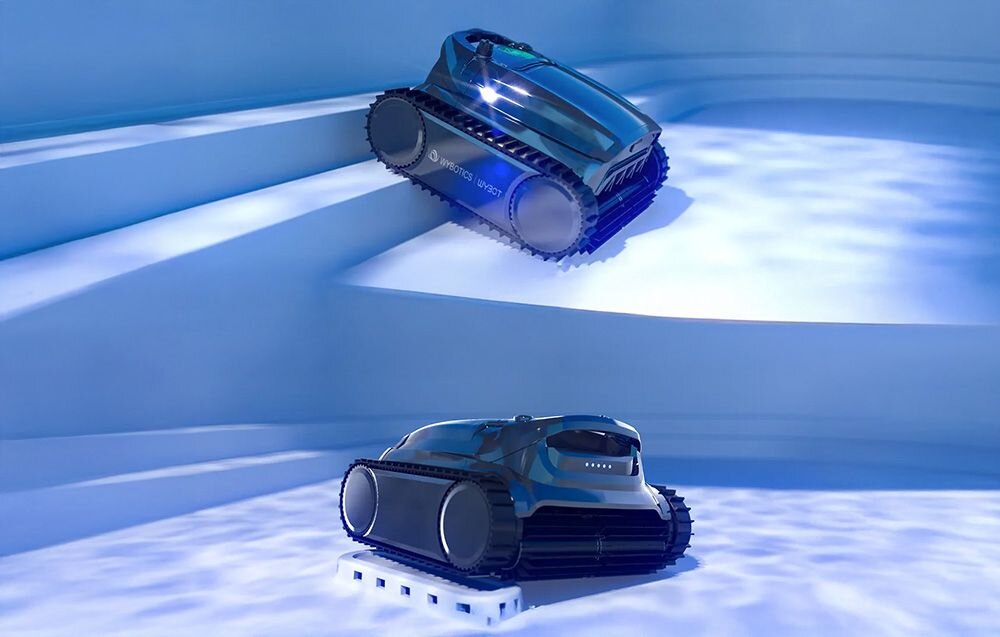 Dwa roboty do czyszczenia basenów znajdują się na schodach basenu, mają ciemne obudowy i gąsienicowe koła. Na jednym z robotów widoczny jest napis \'WYBOT M1\'.