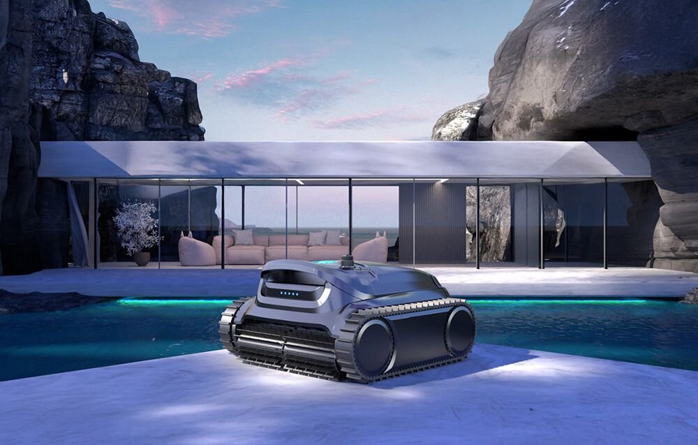 Na pierwszym planie znajduje się robot do czyszczenia basenów o nowoczesnym wyglądzie, umieszczony na brzegu basenu. W tle widać nowoczesny budynek z dużymi przeszklonymi ścianami, otoczony skałami.