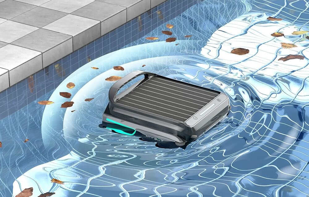 Robot do czyszczenia basenów unosi się na powierzchni wody w basenie, otoczony kilkoma liśćmi. W tle widoczna jest krawędź basenu z płytkami.