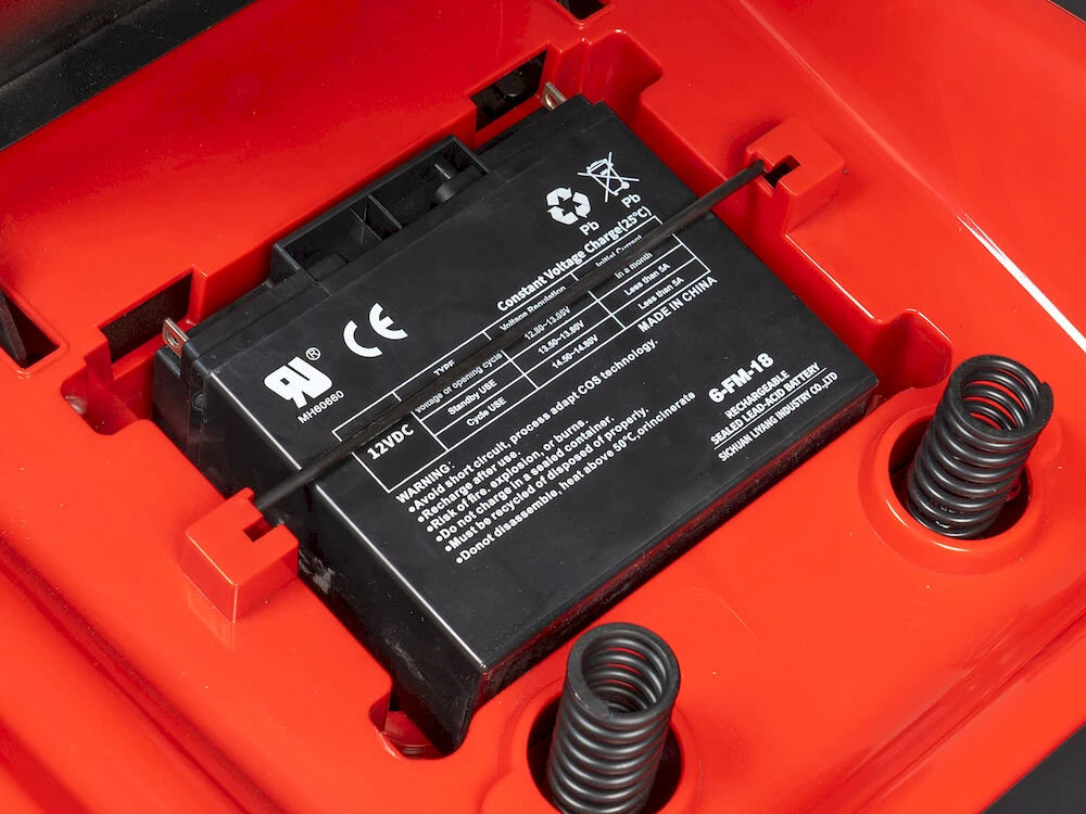 Czarna bateria z oznaczeniami i specyfikacjami technicznymi, umieszczona w czerwonej obudowie. Widoczne są symbole recyklingu i ostrzeżenia dotyczące użytkowania.