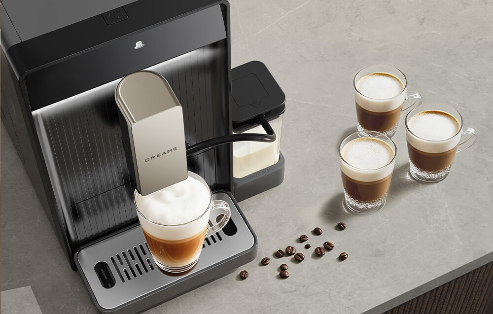 Na blacie kuchennym stoi szary ekspres do kawy z logo \'DREAME\', obok którego znajdują się cztery filiżanki z kawą i spieniacz do mleka. Na blacie leży kilka ziaren kawy.