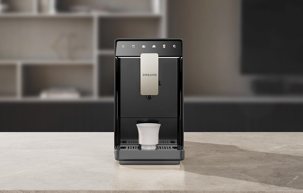 Na blacie stoi szary ekspres do kawy z logo \'DREAME\', z filiżanką umieszczoną pod dyszą. W tle widoczne są rozmyte półki.