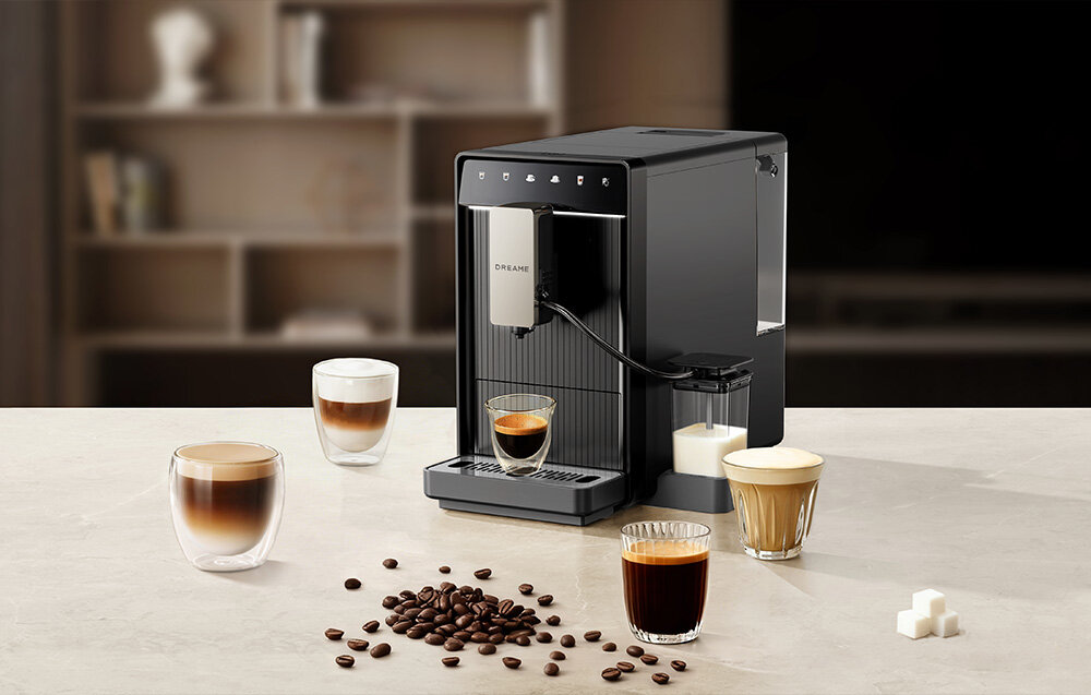 Na blacie stoi szary ekspres do kawy z logo DREAME, obok którego znajdują się filiżanki z różnymi rodzajami kawy oraz rozsypane ziarna kawy. W tle widoczna jest rozmyta półka z książkami.