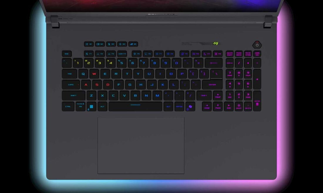Laptop z podświetlaną klawiaturą w różnych kolorach, z dużym touchpadem poniżej. Na obudowie widoczne jest logo producenta oraz przycisk zasilania z podświetleniem.