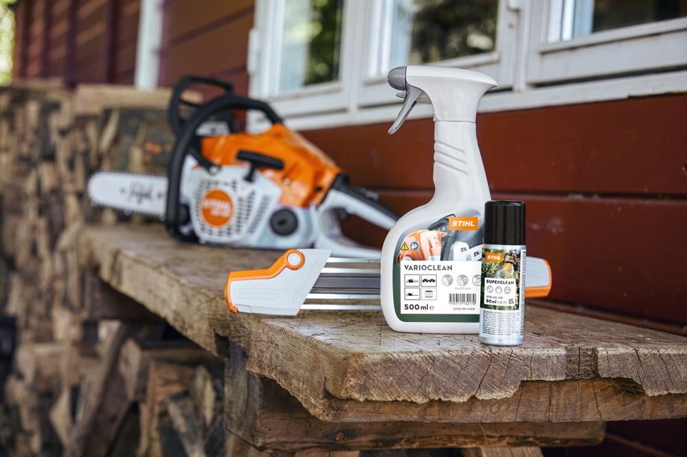 Na drewnianej ławce znajduje się butelka środka czyszczącego STIHL VariClean 500ml oraz puszka smaru. W tle widoczna jest piła łańcuchowa.