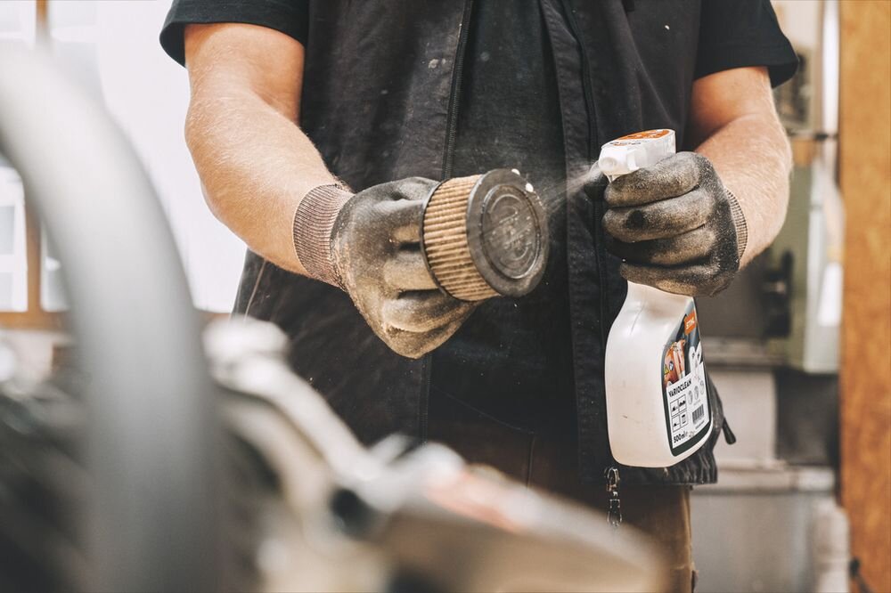 Osoba w rękawicach trzyma butelkę środka czyszczącego i spryskuje filtr powietrza. Na butelce widoczne jest logo STIHL i napis \'VariClean\'.