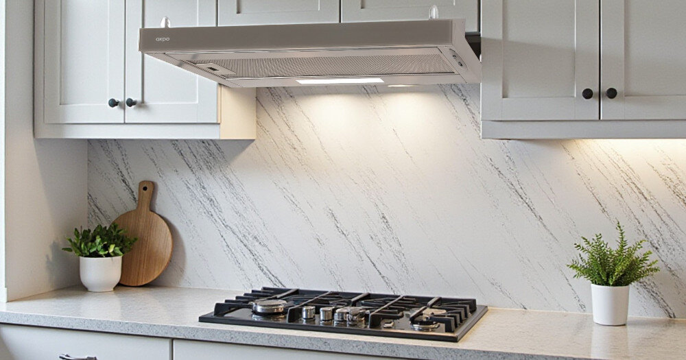 OKAP AKPO WK-7 Light Glass Eco 60 Na zdjęciu 22.jpg widać jasną kuchnię z czarnym, szklanym okapem skośnym zamontowanym nad gazową płytą grzejną z czterema palnikami. Blat zdobi doniczka z rośliną oraz deski do krojenia i owoce, co podkreśla minimalistyczny, elegancki wystrój wnętrza