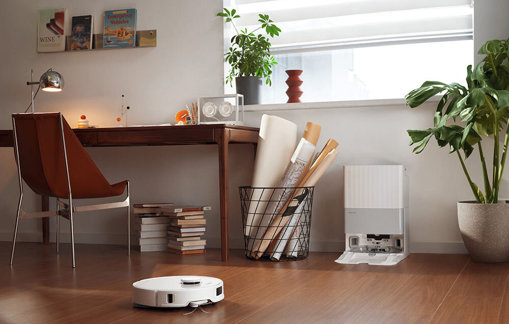 W nowoczesnym wnętrzu z drewnianą podłogą znajduje się robot sprzątający w kolorze białym. Obok biurka z książkami i roślinami stoi stacja dokująca.