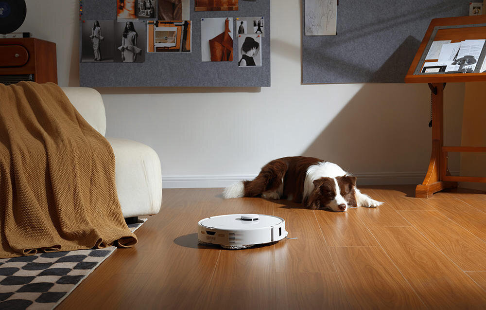 Na drewnianej podłodze znajduje się biały robot sprzątający. W tle leży pies, a na ścianie wiszą różne zdjęcia i grafiki.