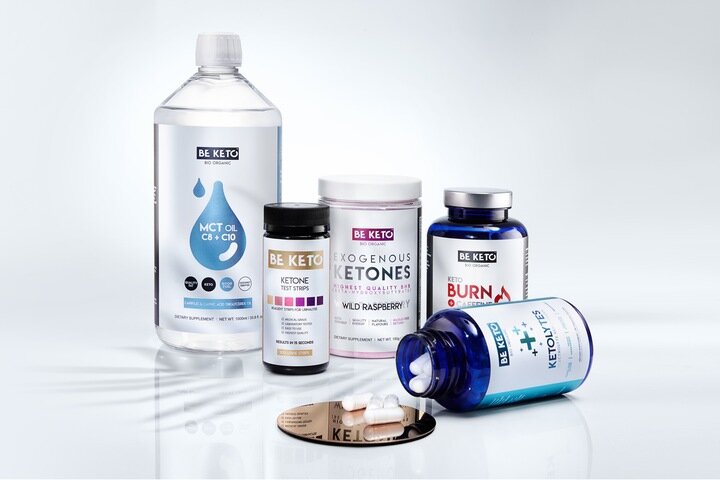 Na obrazie znajduje się zestaw produktów marki BE KETO, w tym butelka oleju MCT, opakowanie egzogennych ketonów o smaku dzikiej maliny, testery ketonowe oraz kapsułki KETO BURN i KETOLYTES. Produkty są ustawione na białym tle, a niektóre z nich są otwarte, ukazując zawartość.