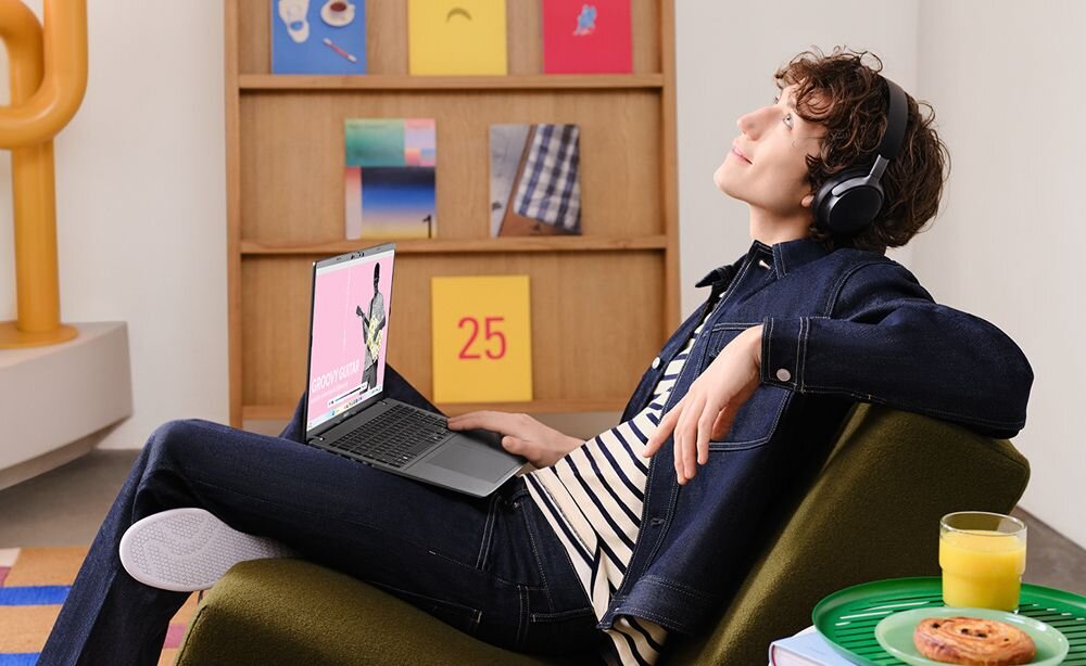 Osoba w słuchawkach relaksuje się na zielonym fotelu, korzystając z laptopa ASUS VivoBook S 14; na ekranie widoczny interfejs aplikacji muzycznej z napisem GROOVY GUITAR i sylwetką gitarzysty na różowym tle, w tle regał z kolorowymi grafikami, na stoliku obok szklanka soku pomarańczowego i talerz z ciastkiem.   - Wzmocnione wrażenia audio Dolby Atmos