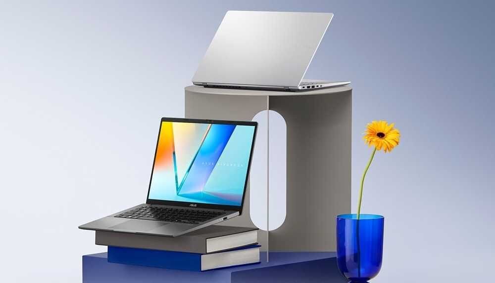 Dwa laptopy ASUS Vivobook S14 – jeden otwarty z kolorowym ekranem ustawiony na stosie książek, drugi częściowo zamknięty na podwyższeniu, obok niebieski wazon z żółtym kwiatem, całość na jasnym, minimalistycznym tle.      