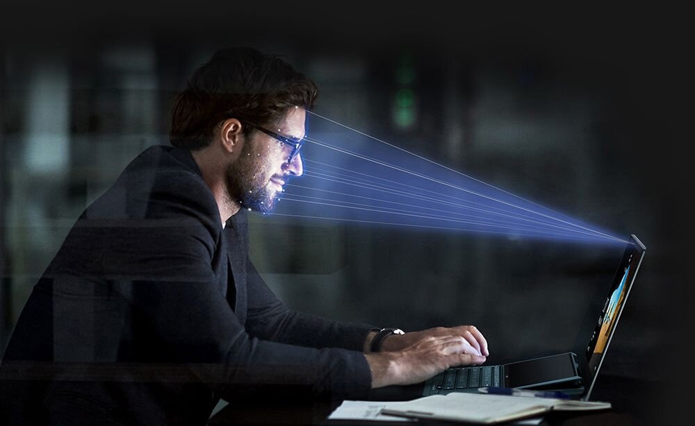 Mężczyzna w ciemnej marynarce siedzi przy biurku i korzysta z laptopa ASUS Vivobook S14; z laptopa w stronę jego twarzy biegną niebieskie linie symbolizujące skanowanie biometryczne lub rozpoznawanie twarzy, na biurku leży notes i długopis, w tle rozmyte wnętrze biura. - Kamera FHD na podczerwień (IR) z obsługą Windows Hello 