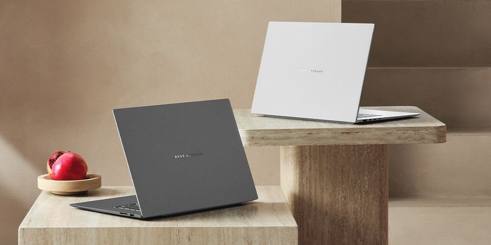 Dwa laptopy stoją na kamiennych stolikach, jeden w kolorze ciemnym, drugi jasnym. Na ciemnym laptopie widoczne jest logo \'ASUS Zenbook\'.