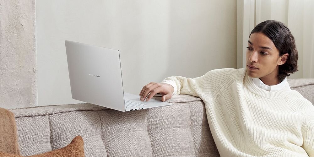 Osoba siedzi na kanapie, obok niej znajduje się otwarty laptop w jasnym kolorze. Na pokrywie laptopa widoczny jest napis \'ASUS Zenbook\'.