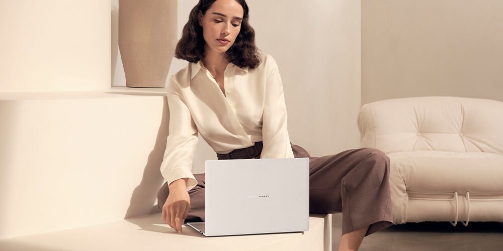 Osoba siedzi na jasnym tle, trzymając biały laptop z widocznym logo producenta. W tle znajduje się jasna sofa.