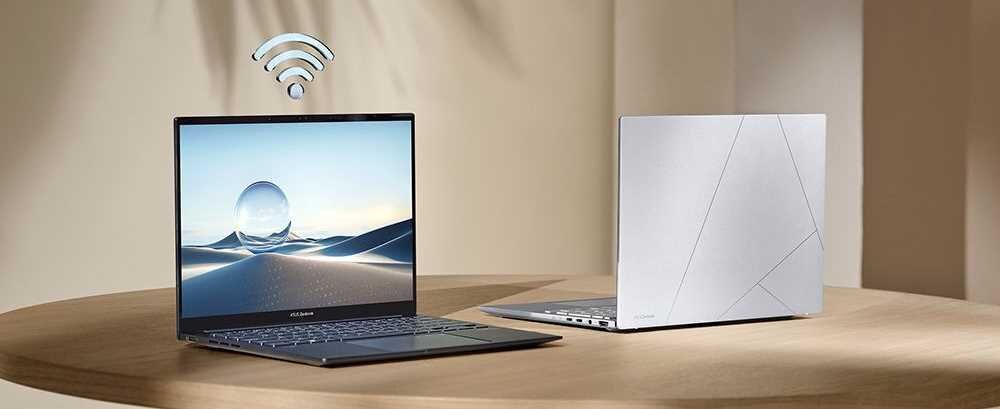 Na drewnianym stole znajdują się dwa laptopy ASUS Zenbook. Jeden z nich jest otwarty, z widocznym ekranem przedstawiającym krajobraz z kulą, a nad nim unosi się symbol Wi-Fi.