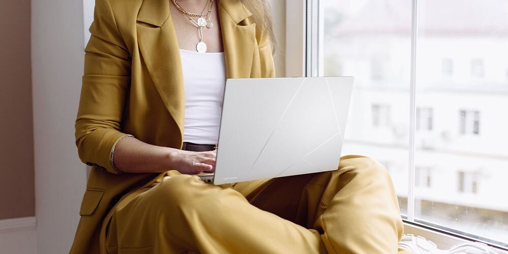 Osoba siedzi przy oknie z laptopem na kolanach. Laptop ma jasną obudowę z subtelnymi liniami na pokrywie.